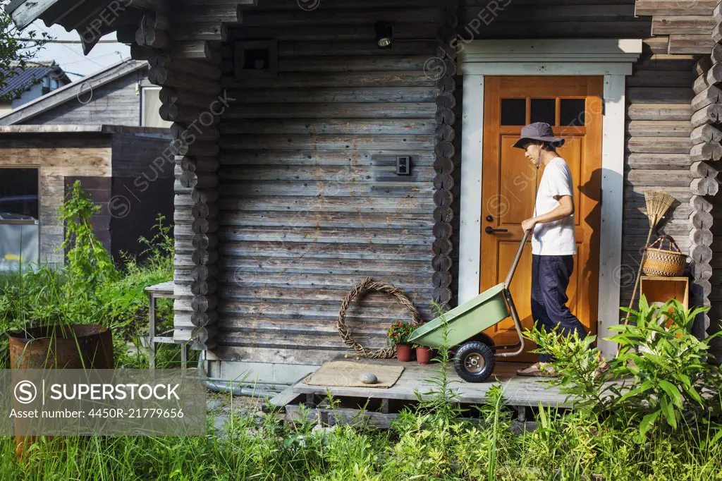Man wearing hat standing outside wooden garden shed, pushing green wheelbarrow.