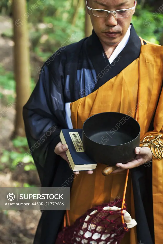 Buddhist monk with shaved head wearing black and yellow robe, standing outdoors, holding prayer book and singing bowl.