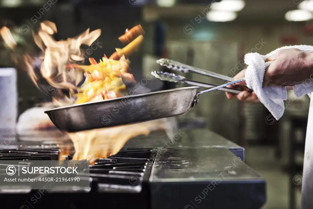 Close-up of chef's hands holding a sauté pan to cook food, flambéing contents. Flames rising from the pan. 