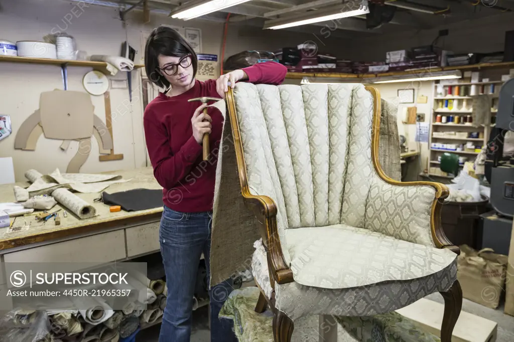 A young female upholsterer using a tack hammer on a chair in an upholstery shop.