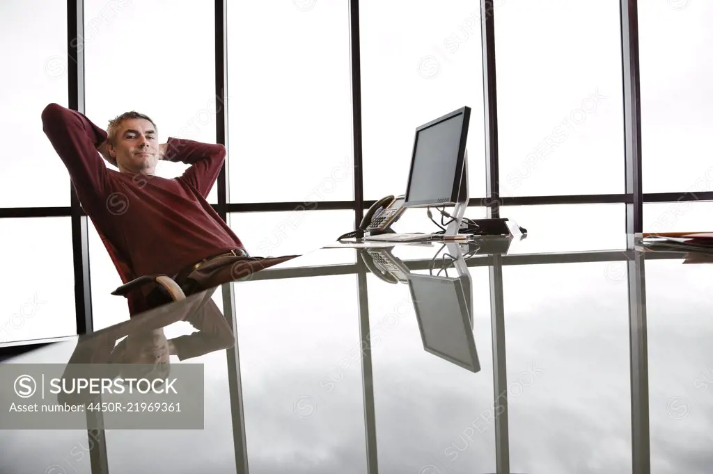 Casually dressed Caucasian businessman relaxed at his reflective desk in front of a large bank of windows.