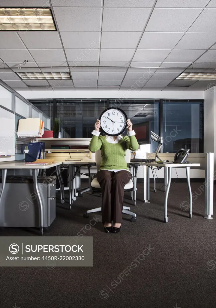 Woman holding a clock face while sitting in her cubicle office space.