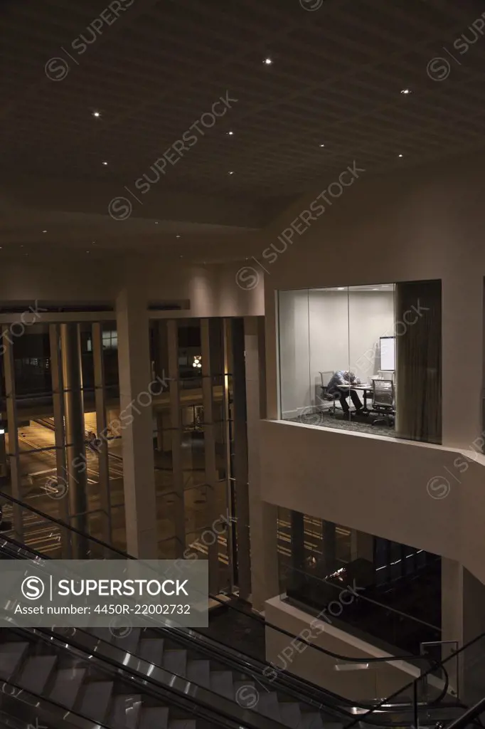 A view looking into a conference room at night  with a single businessman at a conference table.