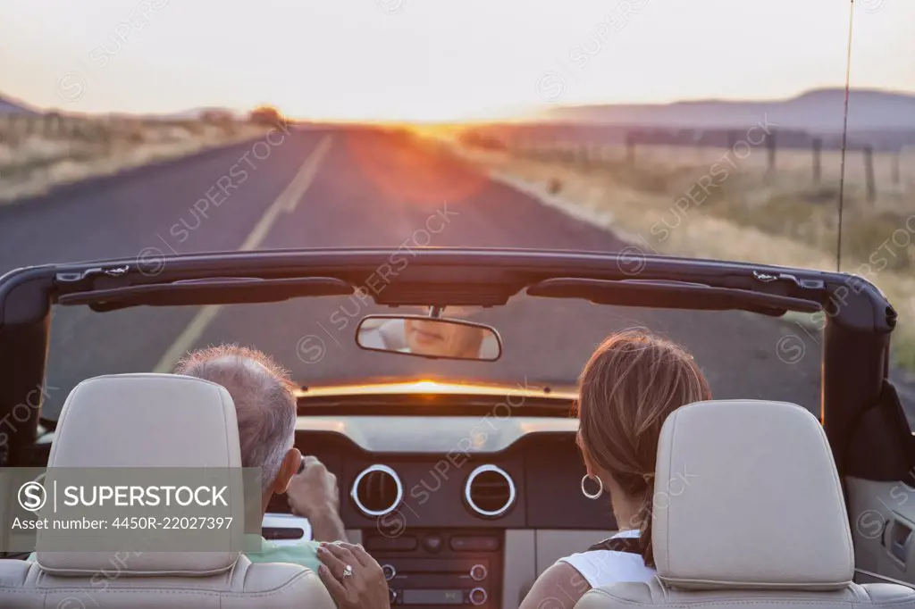View from behind of senior couple in a convertible sports car driving on a highway at sunset in eastern Washington State, USA.