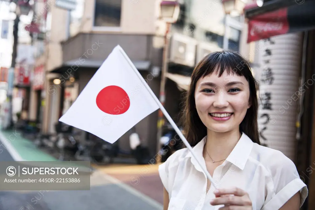 Smiling Japanese woman with long brown hair wearing white short-sleeved blouse standing in a street, holding small Japanese flag.