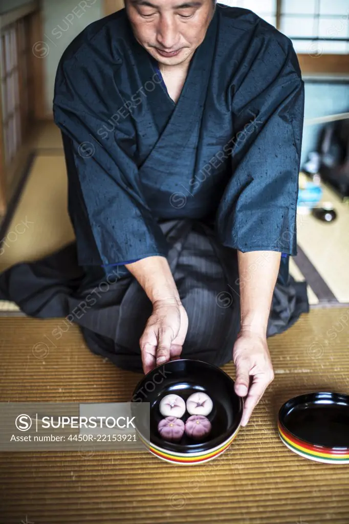 Japanese man wearing traditional kimono knelling on floor, holding a bowl with Wagashi, sweets traditionally served during a Japanese Tea Ceremony.