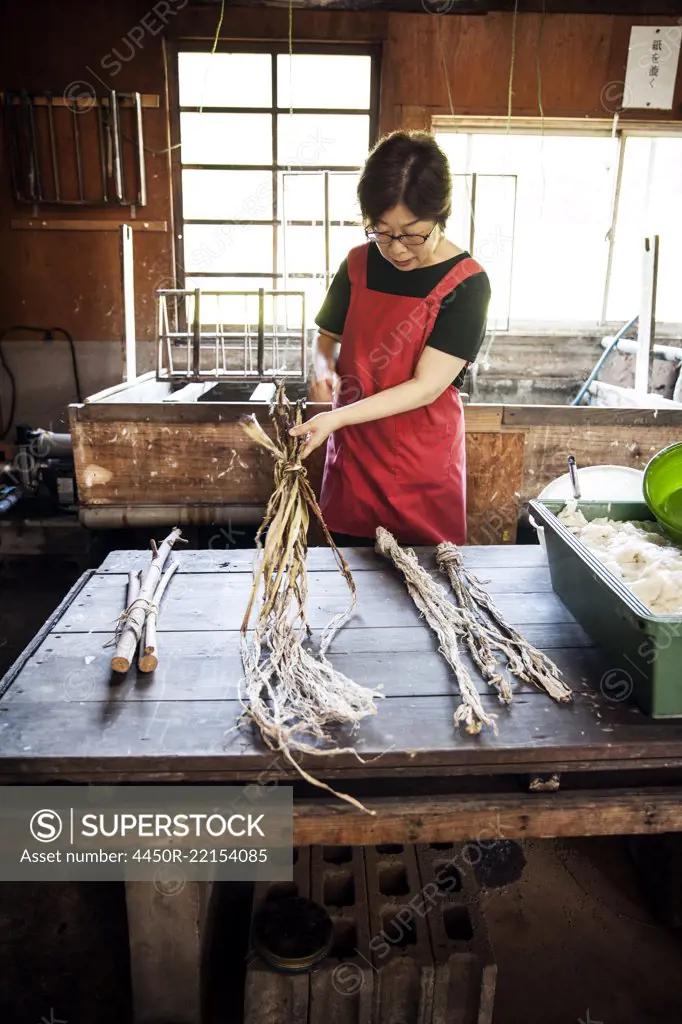 Woman standing at a table in a traditional Washi workshop, stripping bark and fibre off twigs and branches of plant material 