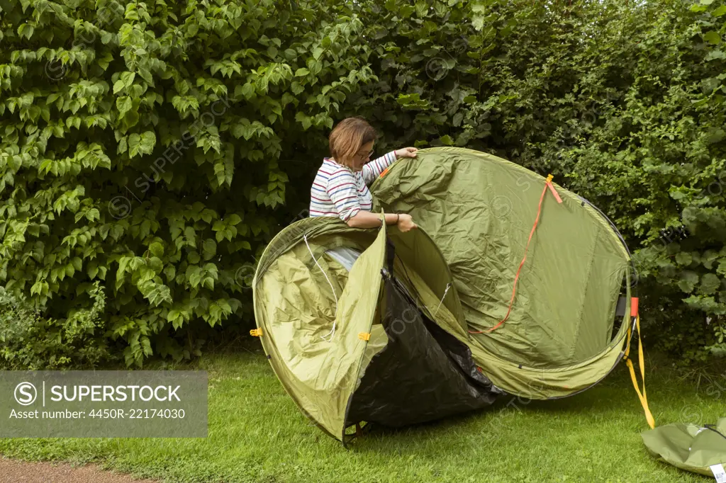 A woman putting up a green pop up tent in a sheltered spot by a tall hedge.