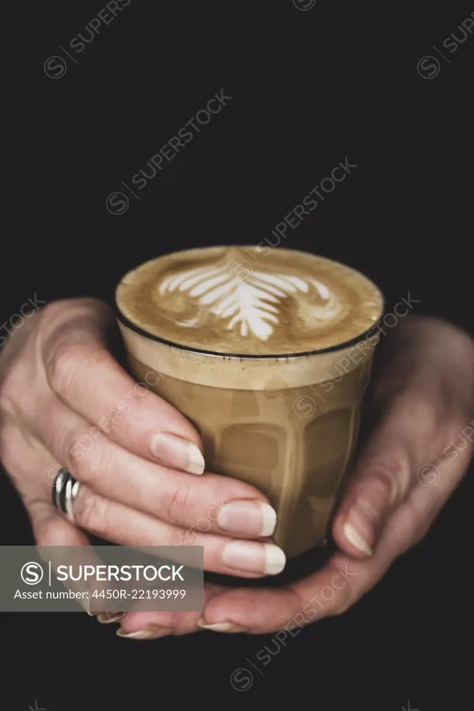 High angle close up of person holding glass with cafe latte topped with intricate foam pattern.