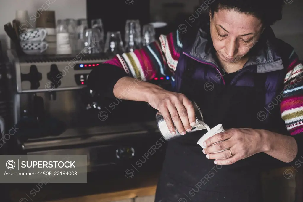 Woman behind espresso machine pouring hot milk into paper cup.