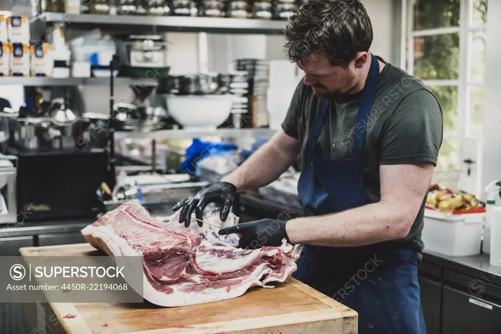 Male butcher wearing apron and black rubber gloves cutting pork ribs on butcher's block.