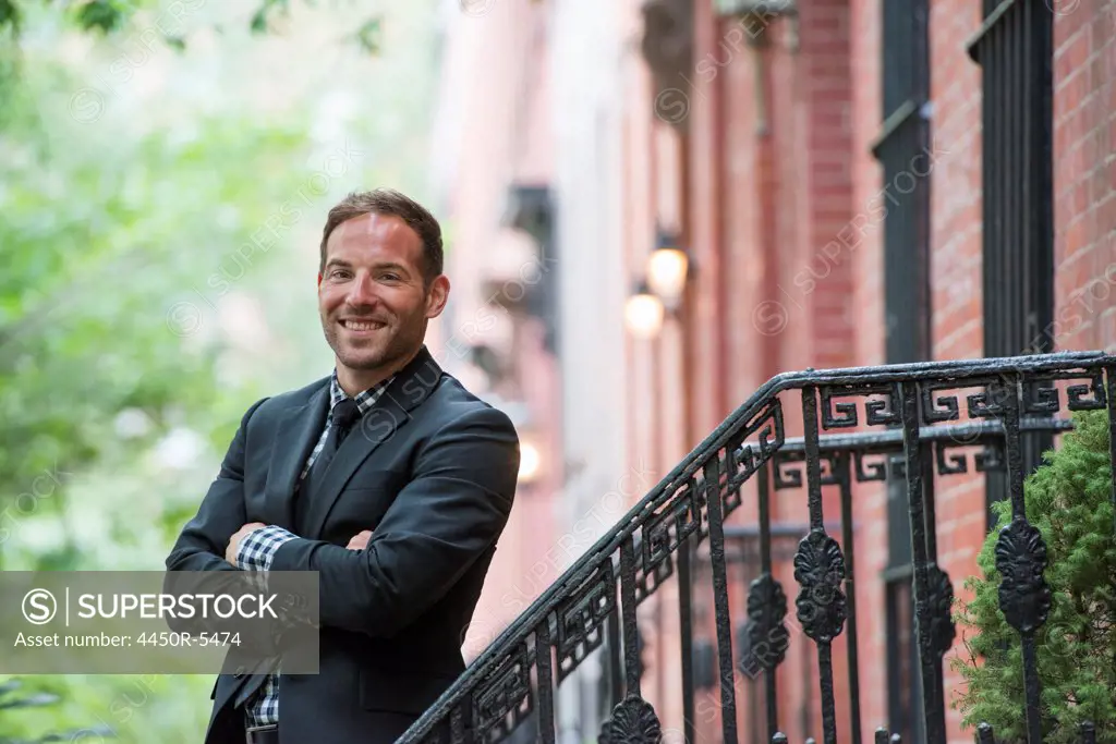 Business people out and about in the city. A man in a suit on the steps of a brownstone building. Arms folded.
