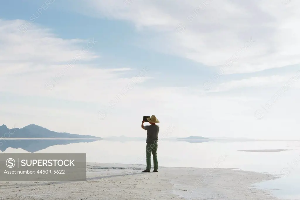 A man standing at edge of the flooded Bonneville Salt Flats at dusk, taking a photograph with a tablet device.