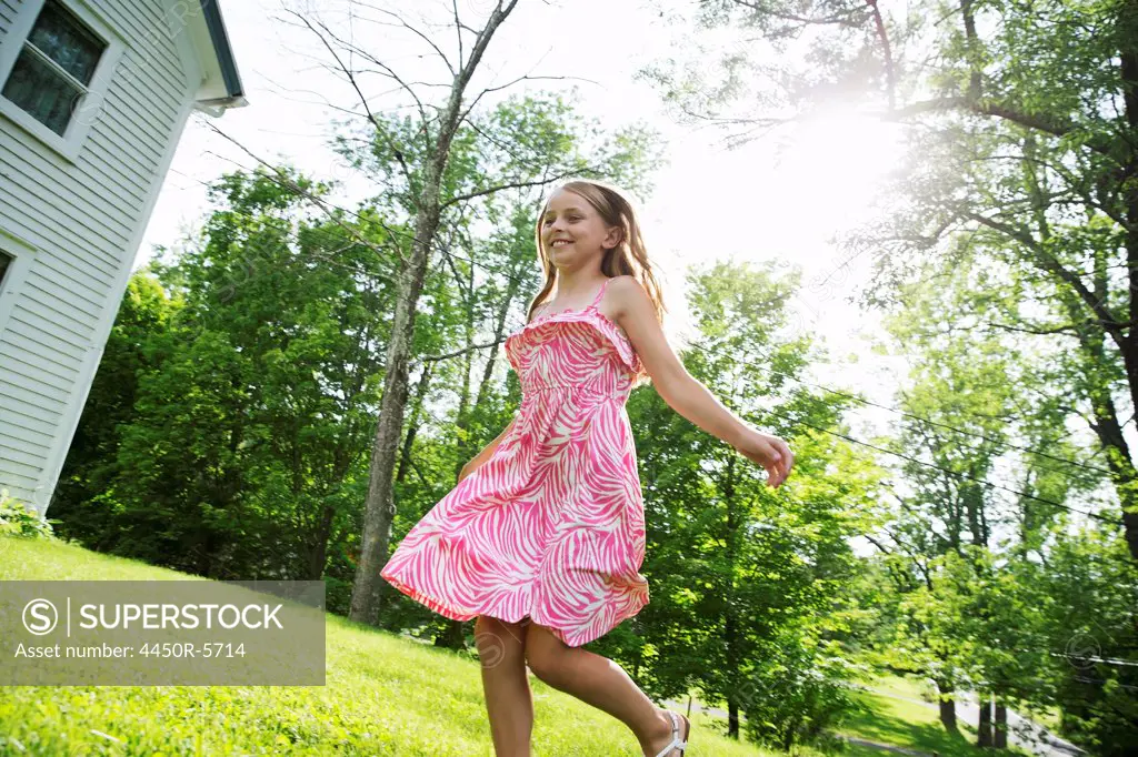 A young girl in a pink patterned sundress running across the grass under the trees in a farmhouse garden.