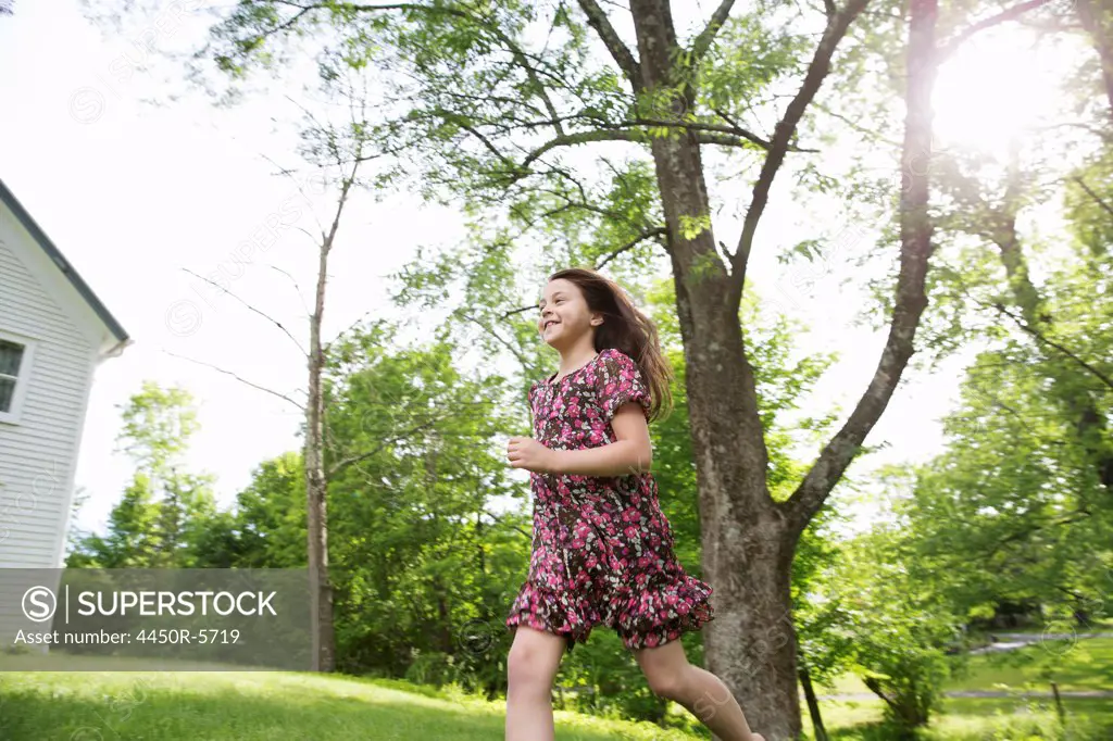 A young girl in a patterned summer dress, running across the grass under the shade of trees in a farmhouse garden.