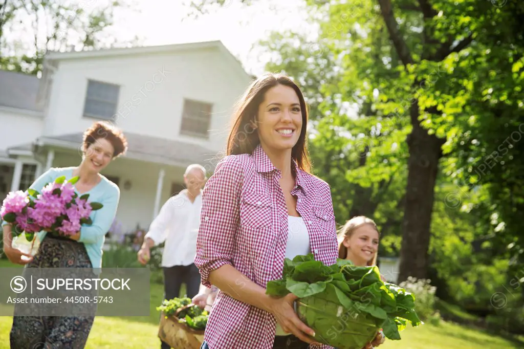 A summer family gathering at a farm. Parents and children walking across the lawn carrying flowers, fresh picked vegetables and fruits. Preparing for a party.