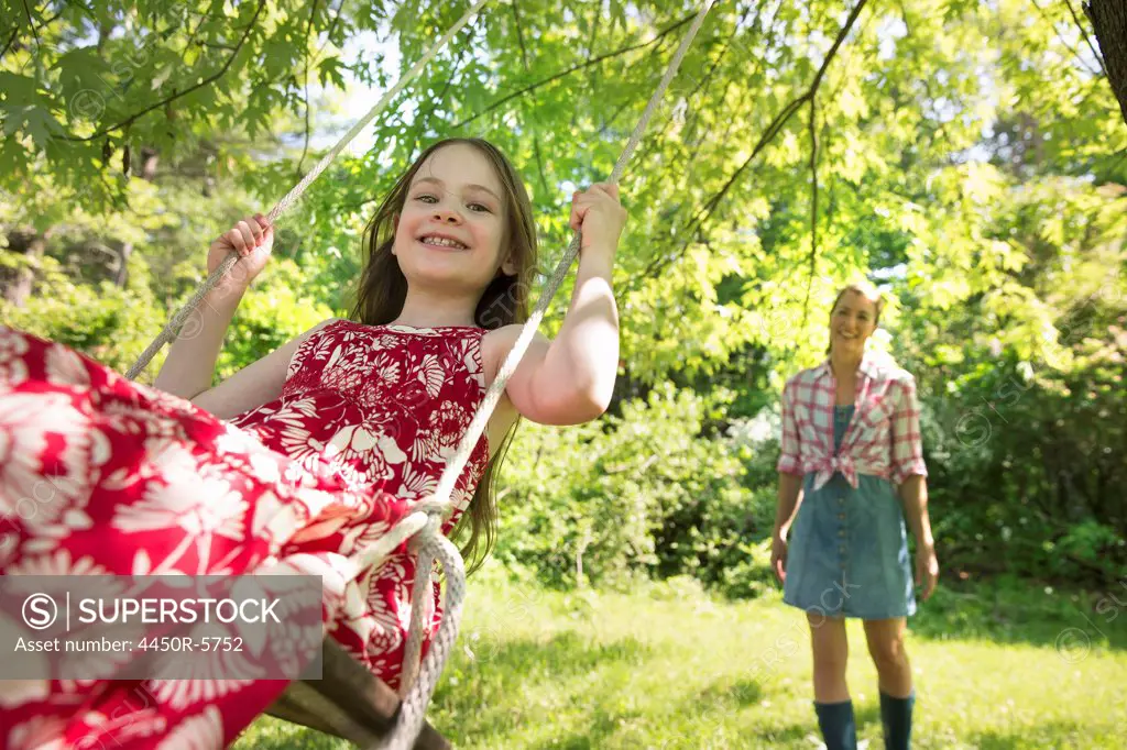 Summer. A girl in a sundress on a swing under a leafy tree. A woman standing behind her.