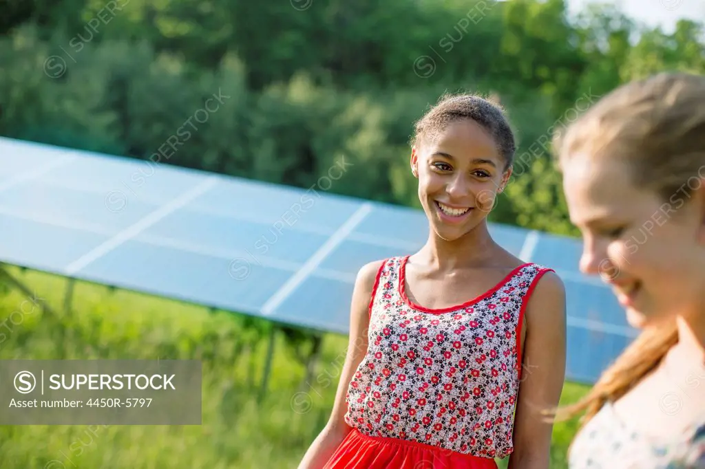 Two young girls on the farm, outdoors. A large solar panel in the field behind them.