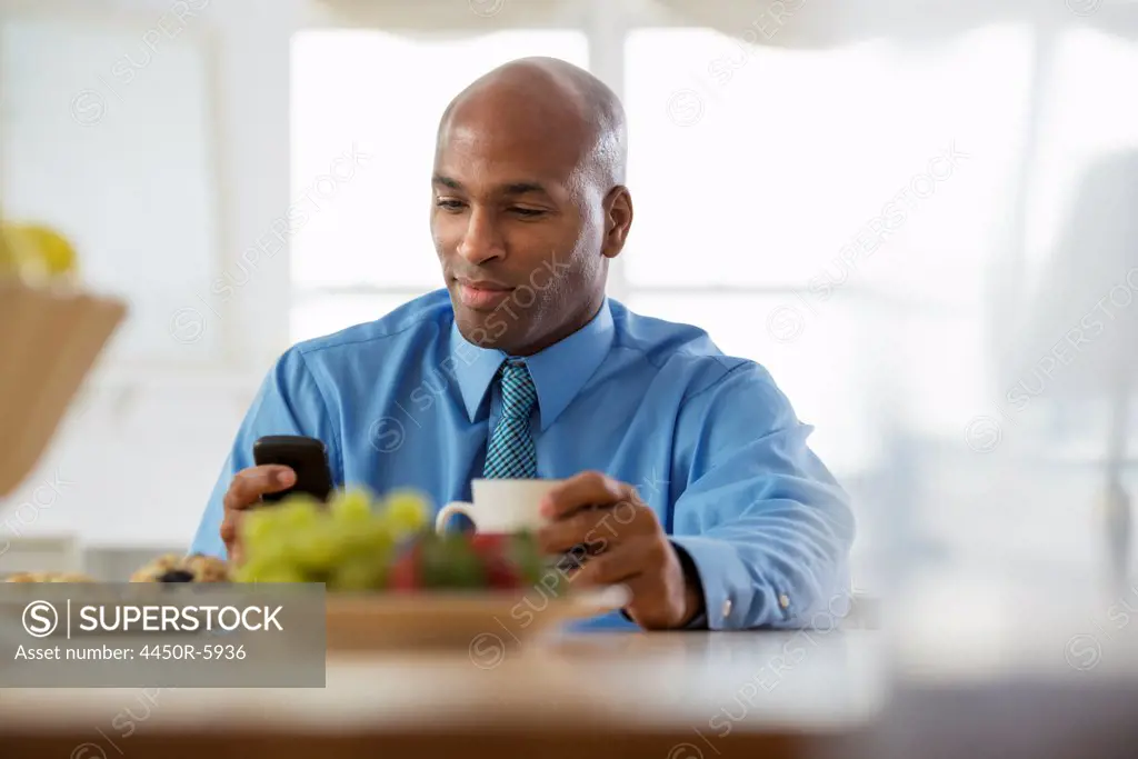 A man in a blue shirt, sitting at a breakfast bar having coffee. A bowl of fruit. Using a smart phone.