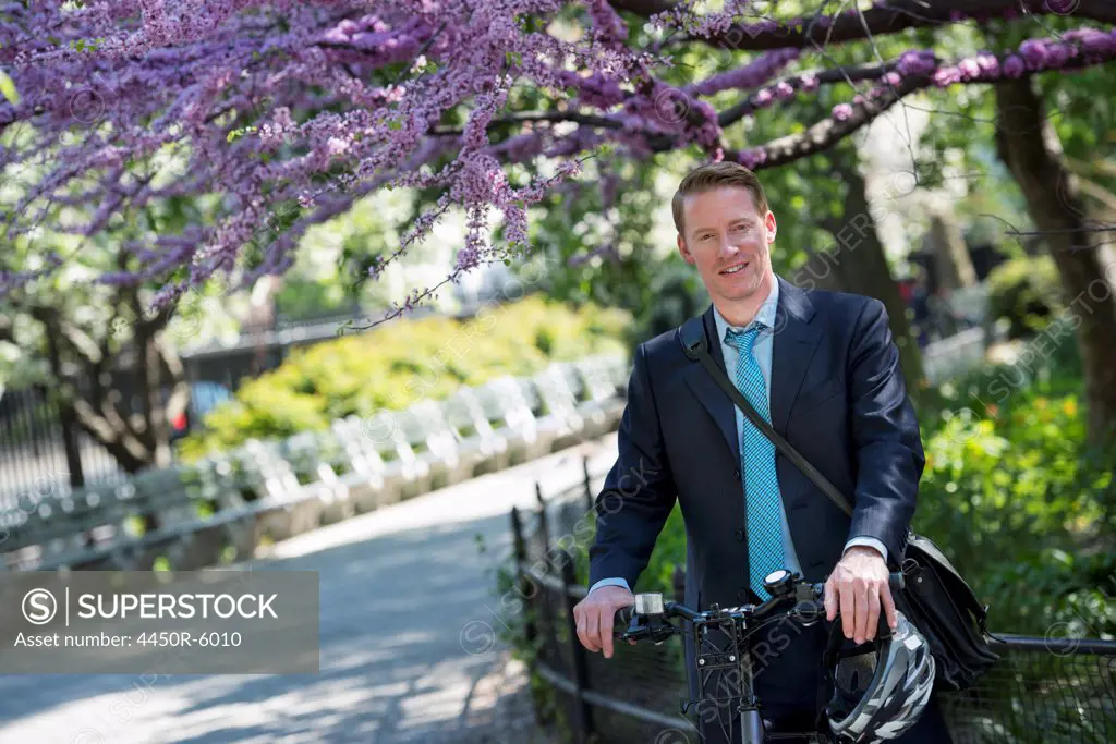 A man in a business suit, outdoors in a park. Sitting on a bicycle, with a cycle helmet in his hands.