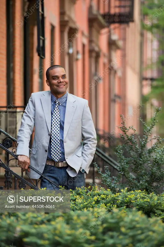 A man in a jacket and tie on a city street outside a terrace of brownstone houses.