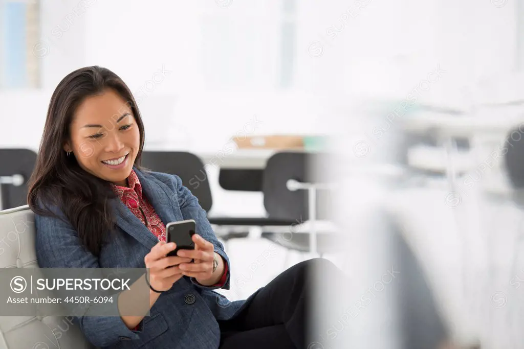 An office in the city. Business. A woman seated in a comfortable chair, checking her smart phone for messages.