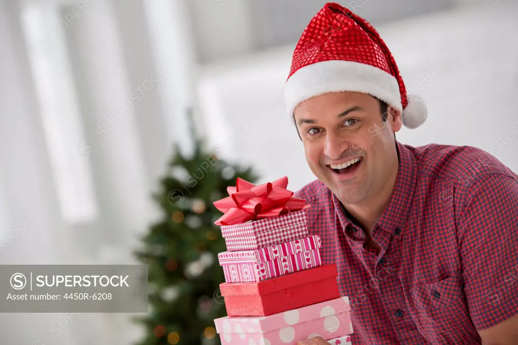 A fresh white building interior, flooded with light. Celebrating Christmas. A man in a Santa hat holding a stack of presents. Decorated Christmas tree.