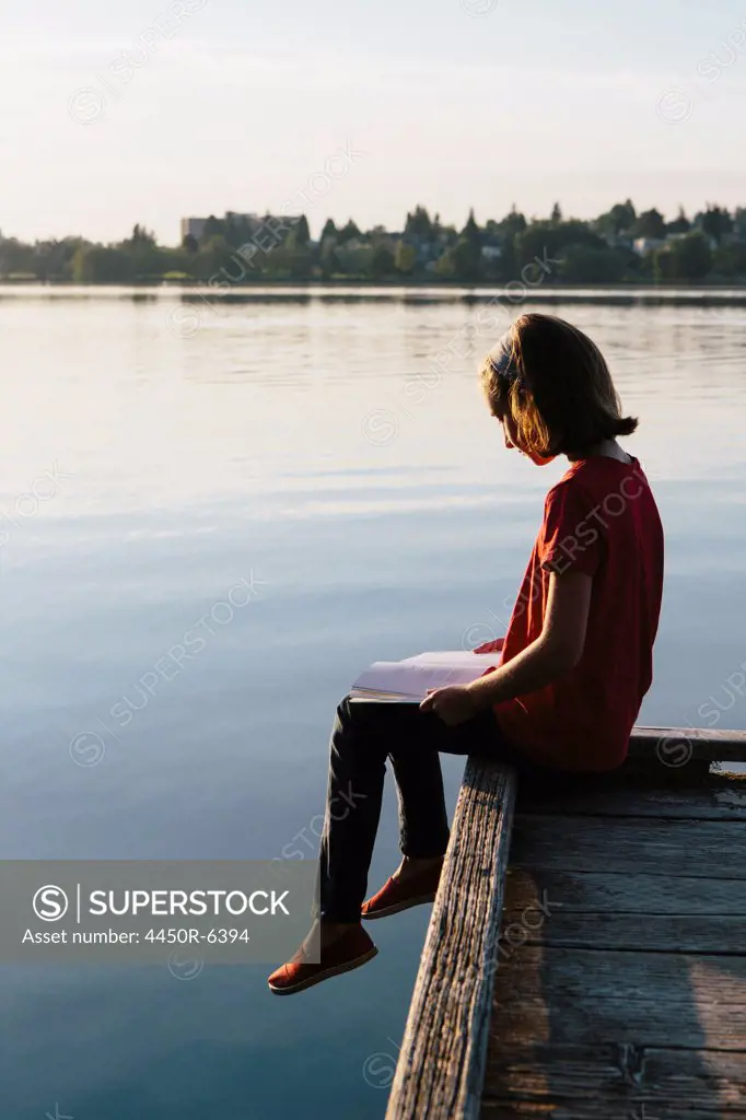 A young girl sitting on a dock, reading a book.
