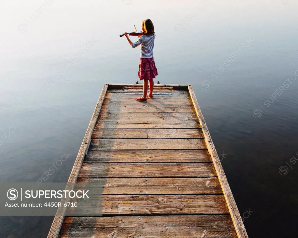 A ten year old girl playing the violin at dawn on a wooden dock.