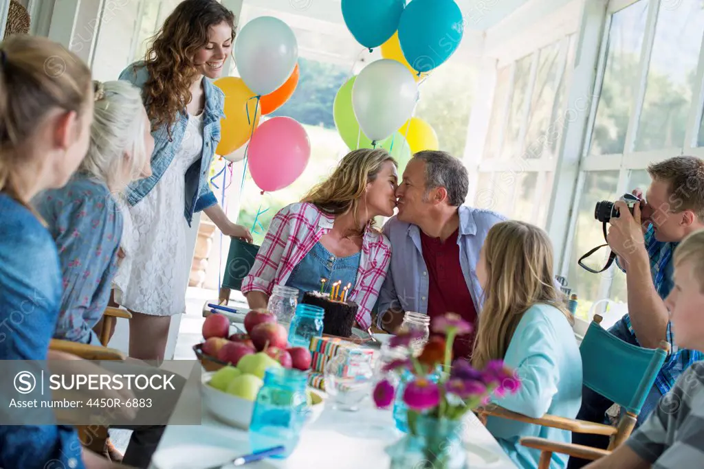 A birthday party in a farmhouse kitchen. A group of adults and children gathered around a chocolate cake.