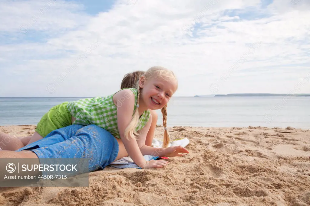 A boy lying on his front on the sand looking out to sea. His sister lying on top of him.