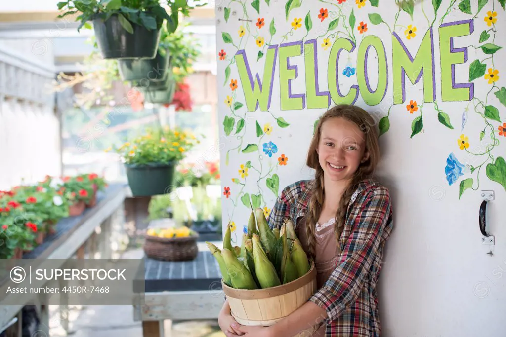 Summer on an organic farm. A girl holding a basket of fresh corn standing by the Welcome sign.