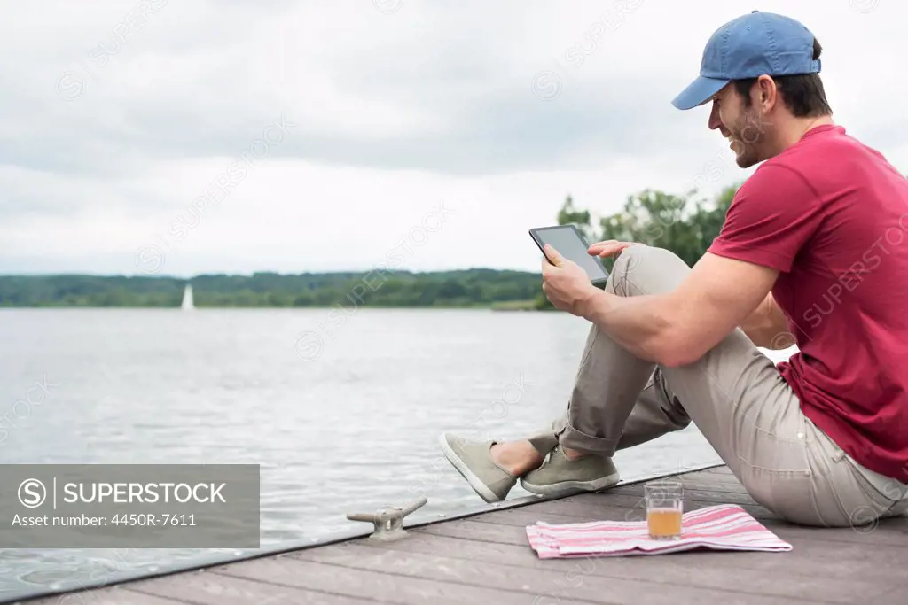 A man seated on a jetty by a lake, using a digital tablet.