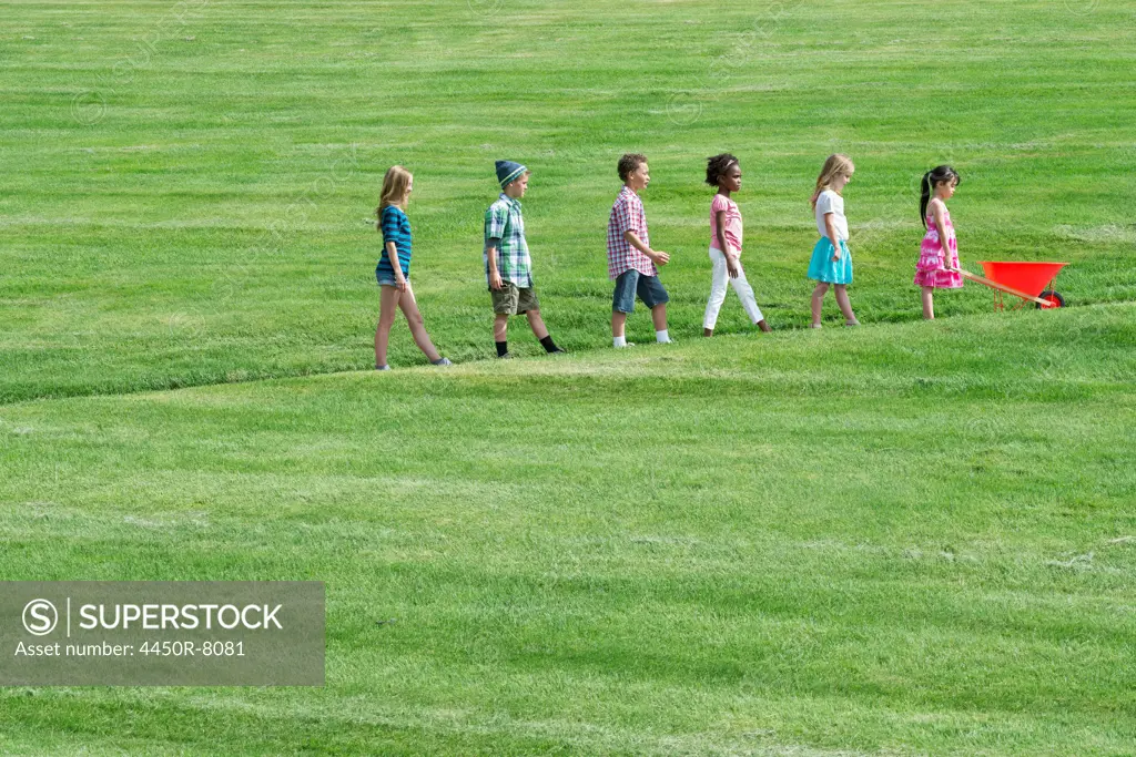 A group of children walking up a sloping path in height order following the smallest one at the front with a red wheelbarrow. Utah, USA. 2/17/2012