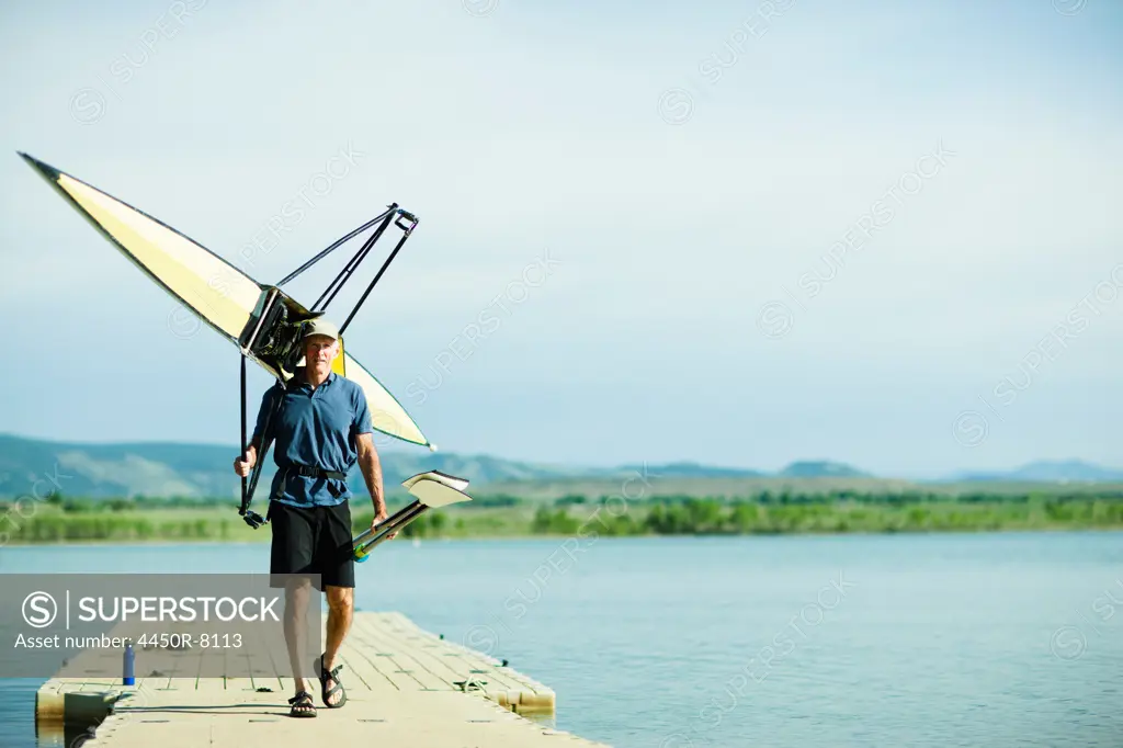 A middle-aged man carrying oars and a rowing shell on his shoulders, on a pontoon. Colorado, USA. 5/23/2012