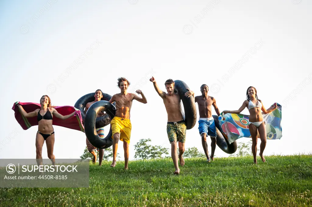 A group of teenagers, boys and girls, running across the grass holding swim towels and inflated floats. Maryland, USA. 7/1/2012