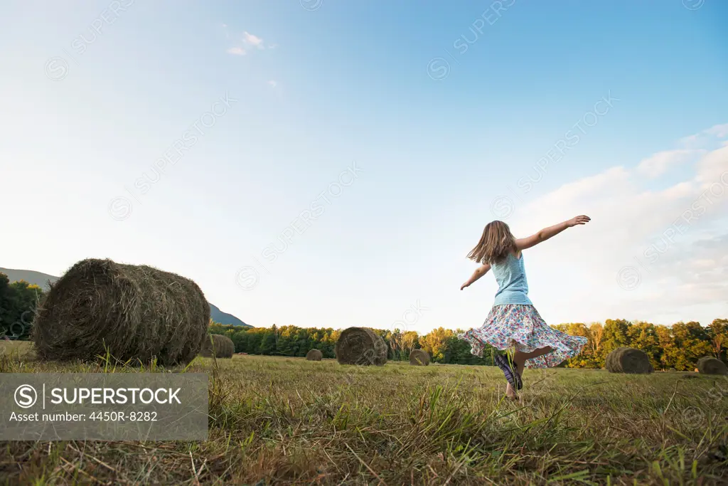 A field full of tall rounded hay bales, and a young girl dancing with her arms outstretched on the stubble field. New York state, USA. 3/21/2012