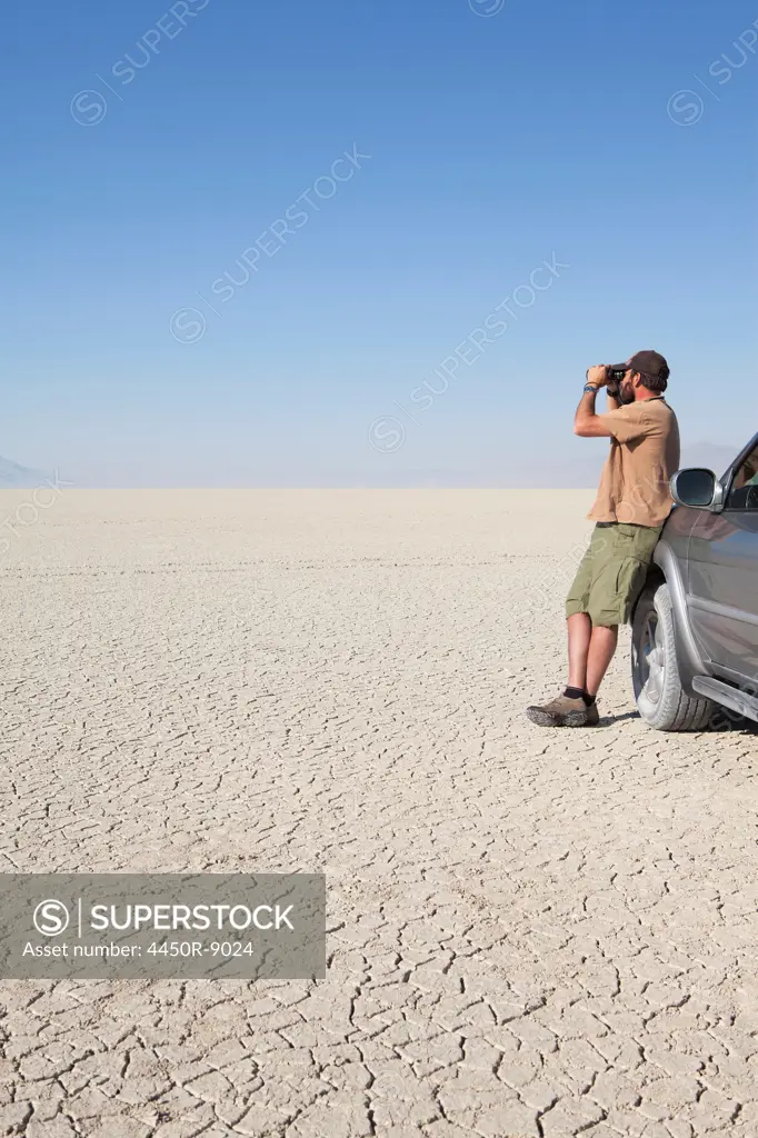 A man standing in a dry desert, looking through binoculars and leaning against a truck, Black Rock Desert, Nevada, USA. 8/8/2012
