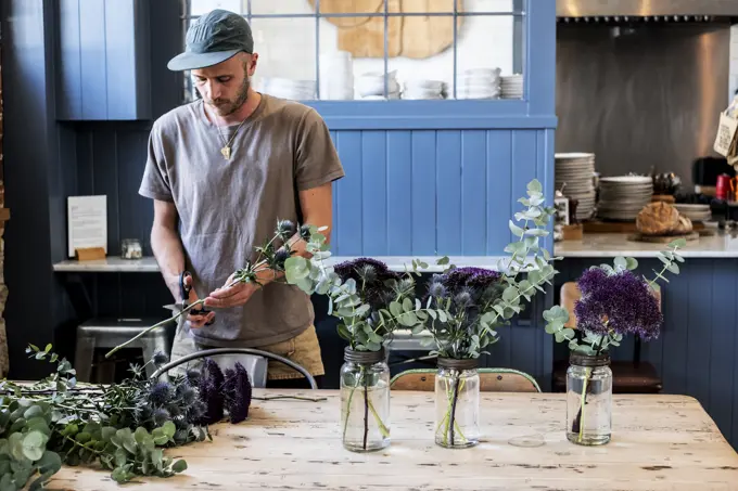 Man wearing baseball cap standing at a table, cutting thistles for flower arrangements in large glass jars.