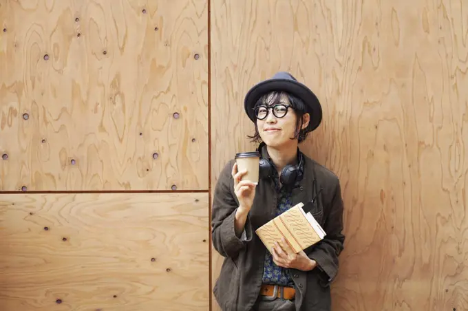 Japanese woman wearing glasses and hat standing outside Eco Cafe, holding paper cup and notebook.