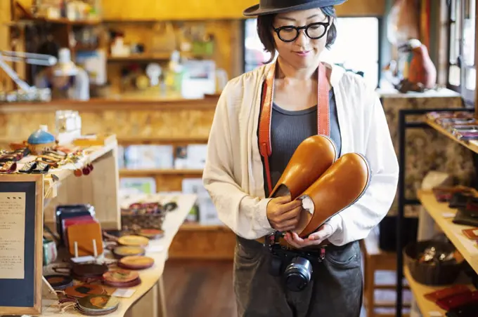 Japanese woman wearing hat and glasses standing in a leather shop, holding pair of leather shoes.