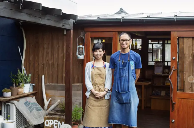 Japanese woman and man wearing blue apron standing outside a leather shop, smiling at camera.
