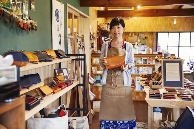 Japanese woman wearing apron standing in a leather shop, holding tan leather clutch bag, smiling at camera.