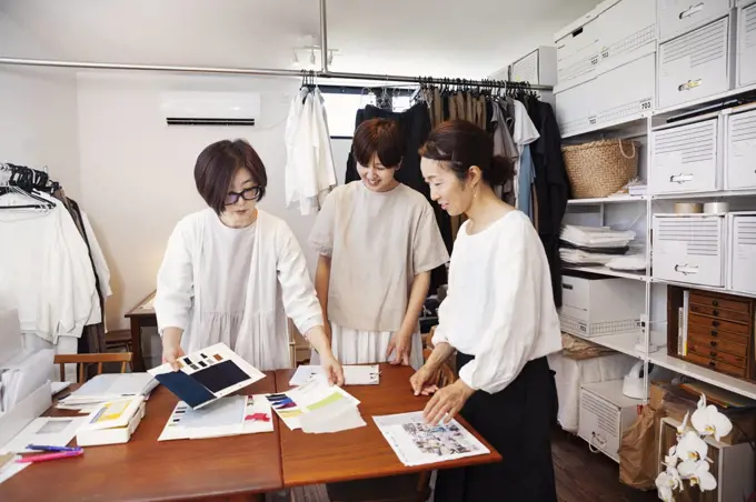 Three Japanese women standing at a table in a small fashion boutique, looking at fabric samples.
