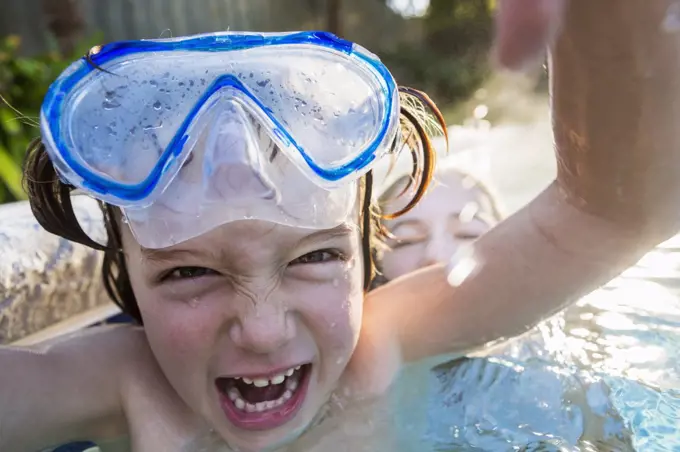 A boy in goggles laughing at the camera, in a warm swimming pool with his sister.