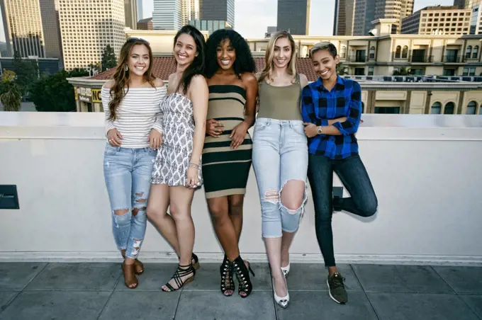 Five young women posing for photographs on a rooftop, city skyline background