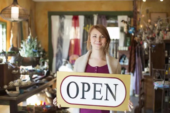 A woman standing in an antique store, holding an OPEN sign. Displays of goods all around her. New York, USA