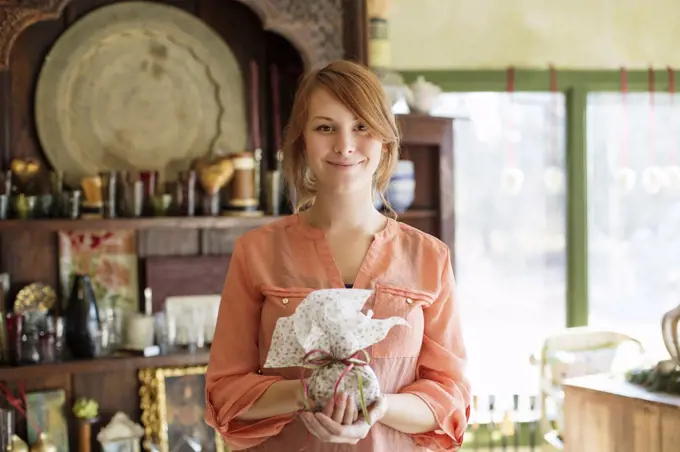 A young woman standing in a store full of antique objects, holding a tied packet with ribbon. New York, USA