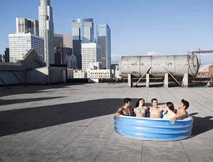 A group of friends, men and women sitting in a small inflatable water pool on a city rooftop, cooling down.