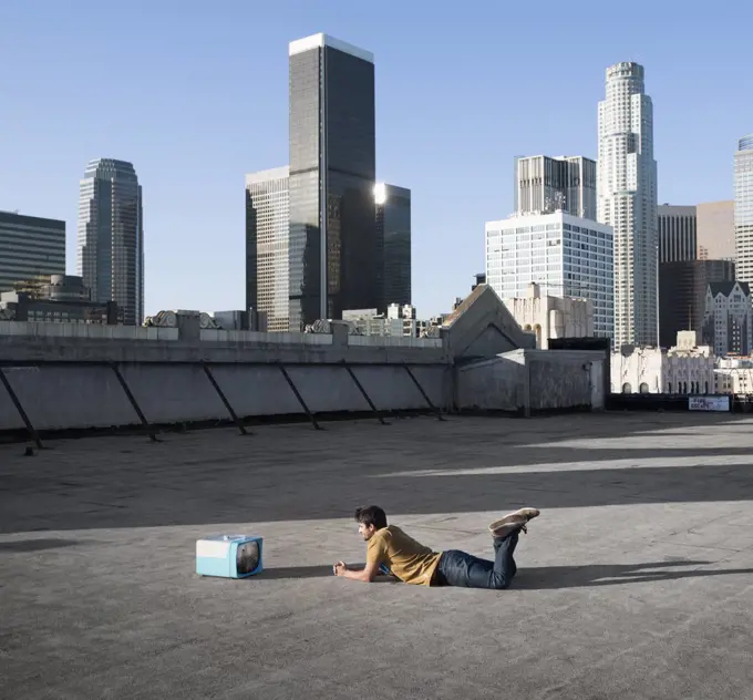 A man lying on his stomach on a city rooftop watching a small blue portable television.